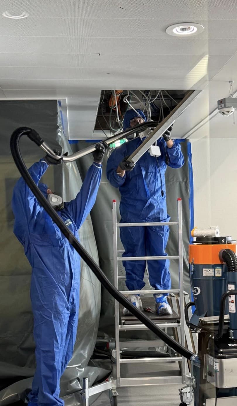 Zwei Mitarbeiter in Schutzanzügen und Halbmasken entfernen Deckendämmung hinter einer Folienabschottung, daneben ein Industriesauger